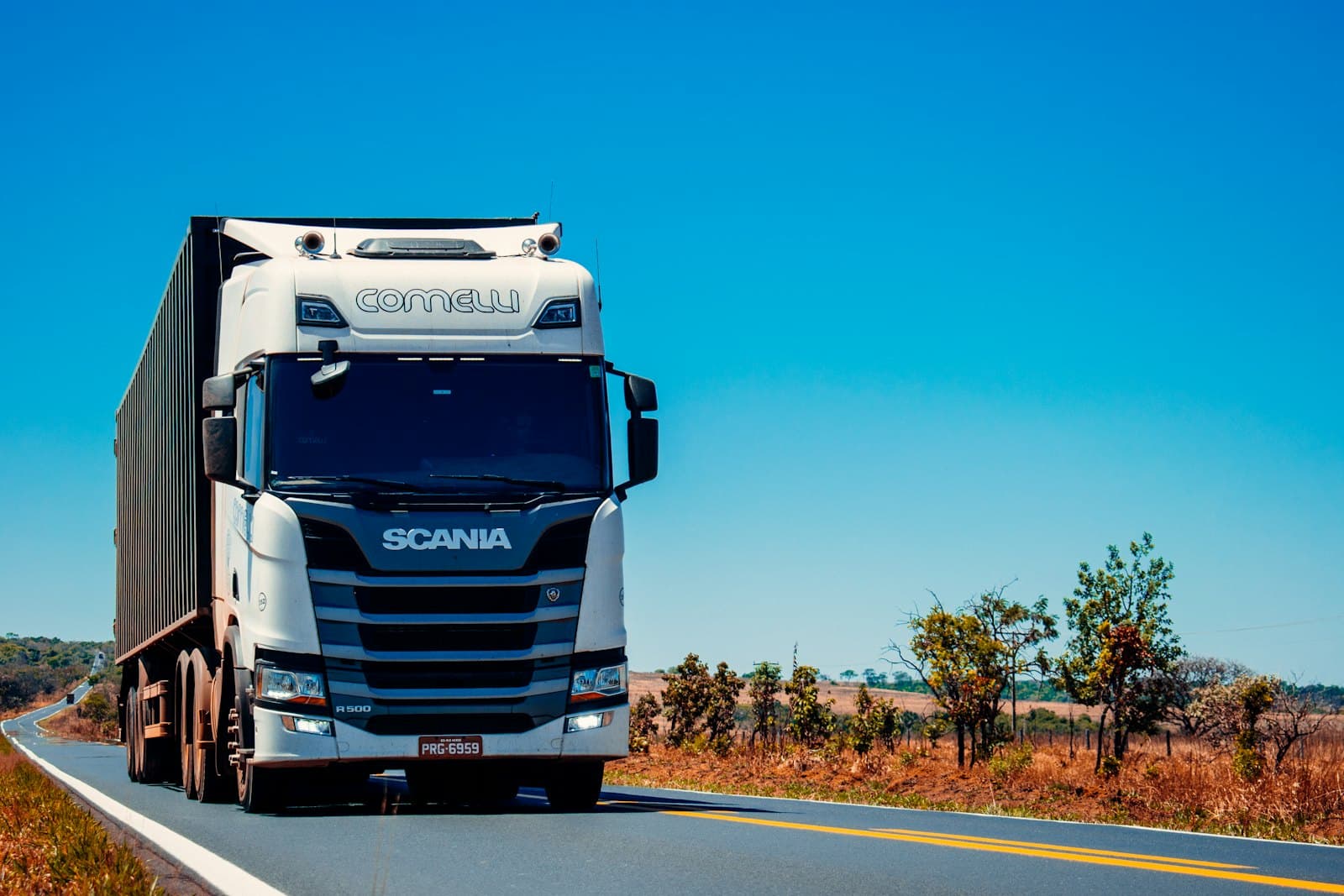 A red conventional semi-truck parked on gravel, photographed from the side.