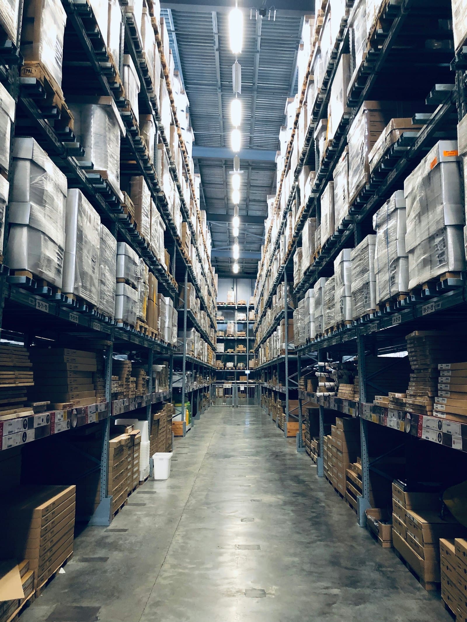 A worker on a forklift in a tall warehouse aisle lined with pallet racking.